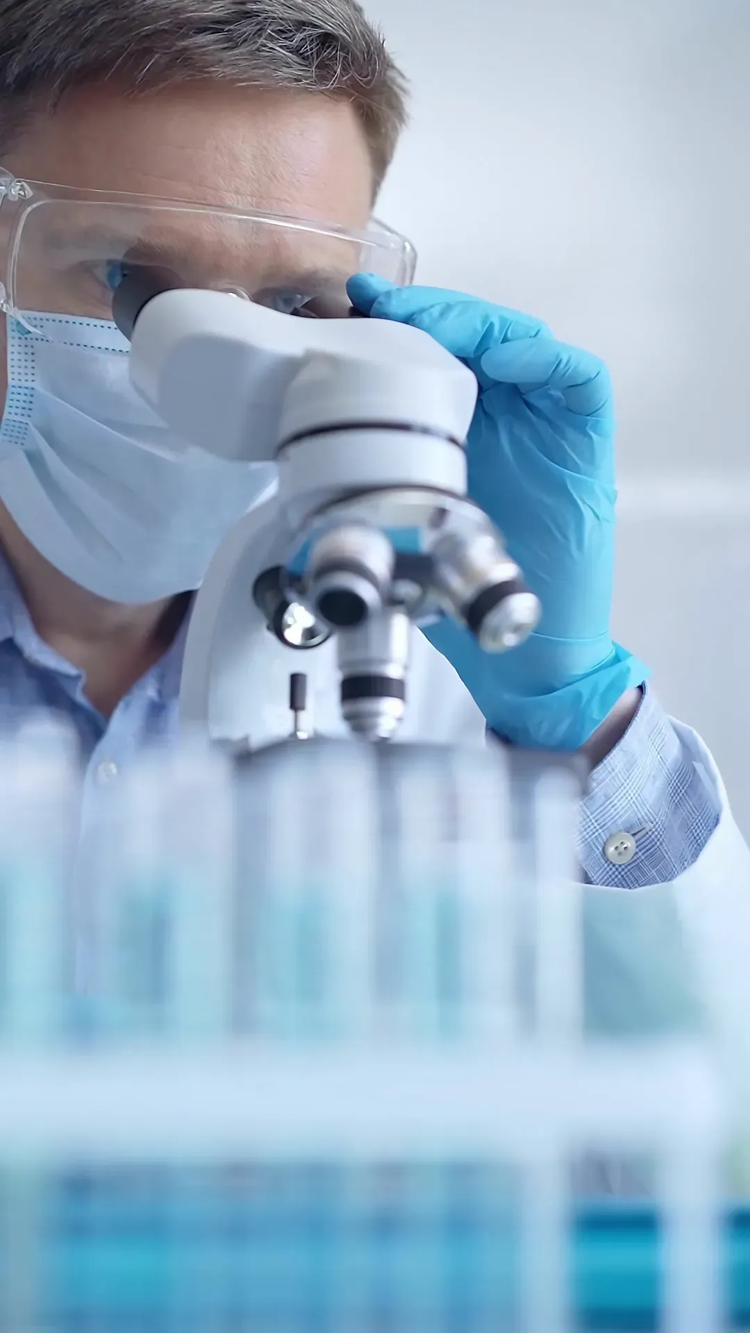 Scientist in safety glasses and blue gloves examining samples through a laboratory microscope